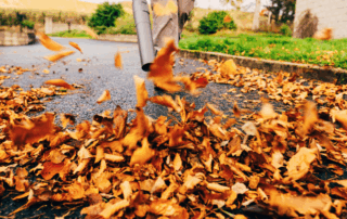 A person using a handheld leaf blower to blast fall leaves from the road